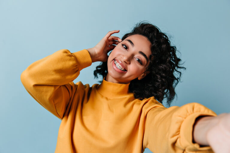 Smiling young woman with traditional metal braces taking a selfie against a blue background