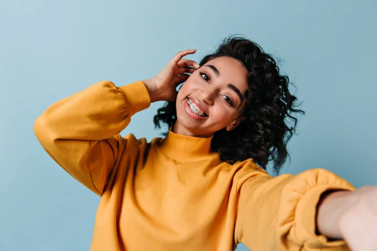 Smiling young woman with traditional metal braces taking a selfie against a blue background